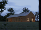 Contemporary barn house with vertical light wood siding and pitched metal roof at dusk, interior lights illuminating bedroom and living spaces