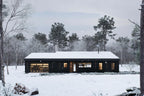 Midcentury modern house plan front elevation with black vertical siding, wood entry and 2-car garage in winter setting