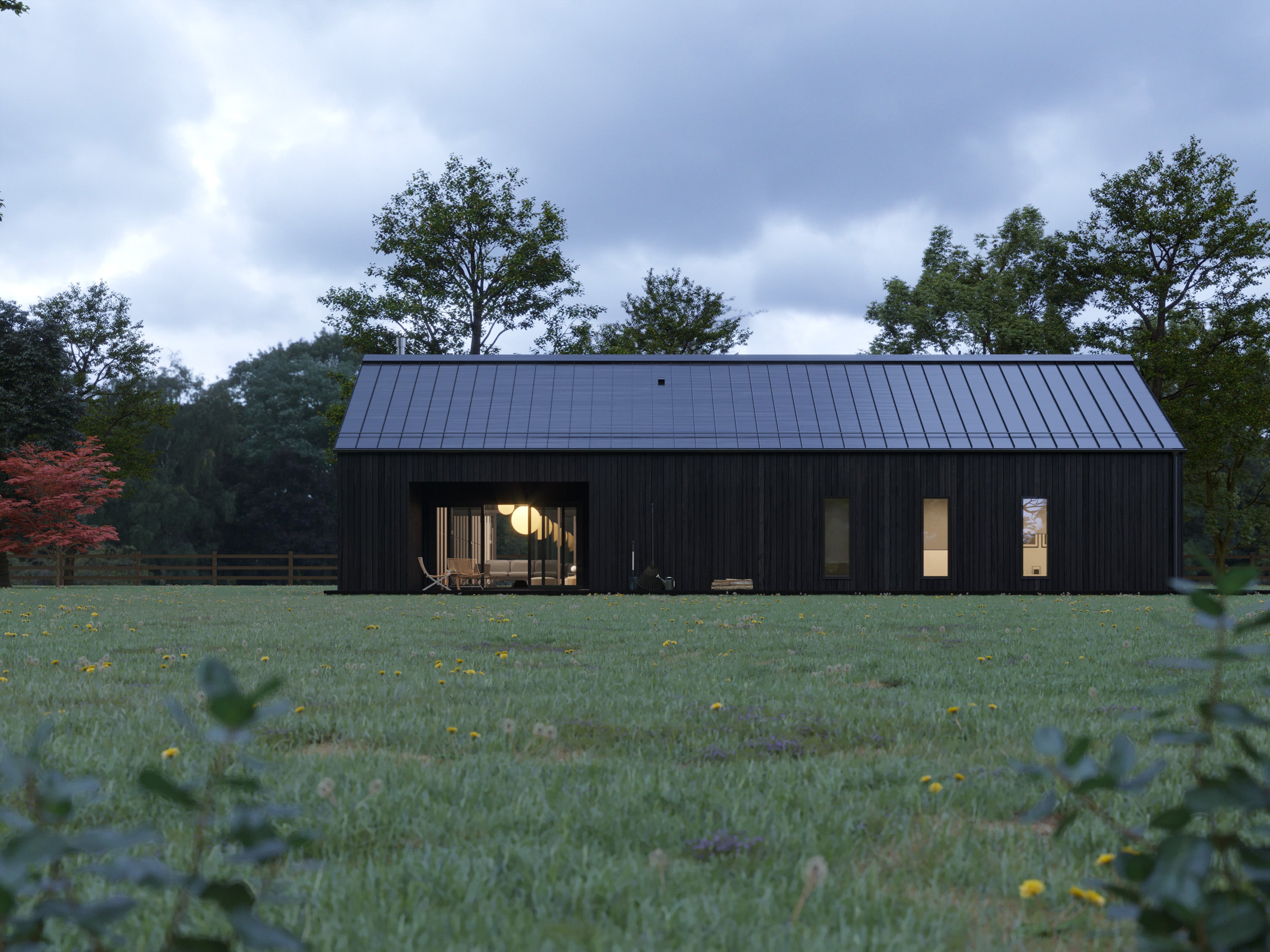 Modern black barn house with charred vertical wood cladding and low-pitch black standing seam metal roof at dusk, warm interior lights glowing through open living area and flanking windows overlooking a wildflower meadow