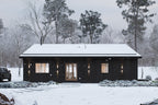 Modern cabin house plan front elevation — black vertical wood siding, wood entry door and sconces in a snowy forest