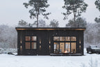 Modern cabin house plan front exterior — black shed-roof cabin with wood siding, deck and fire pit in a snowy forest